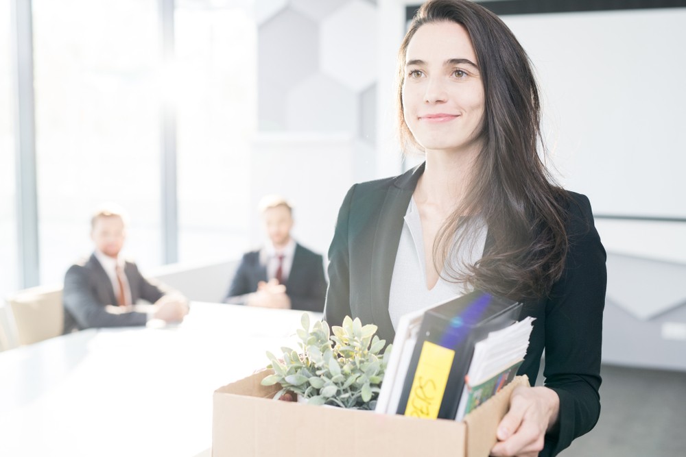 smiling young businesswoman holding box of personal belongings leaving office after quitting job, copy space